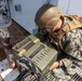 A U.S. Marine in full combat gear is closely inspecting or operating a military equipment case or device on what appears to be the deck of a naval vessel or industrial platform. The Marine is wearing MARPAT digital camouflage, a Kevlar helmet, and a tactical vest with a backpack. A second Marine is partially visible in the background.