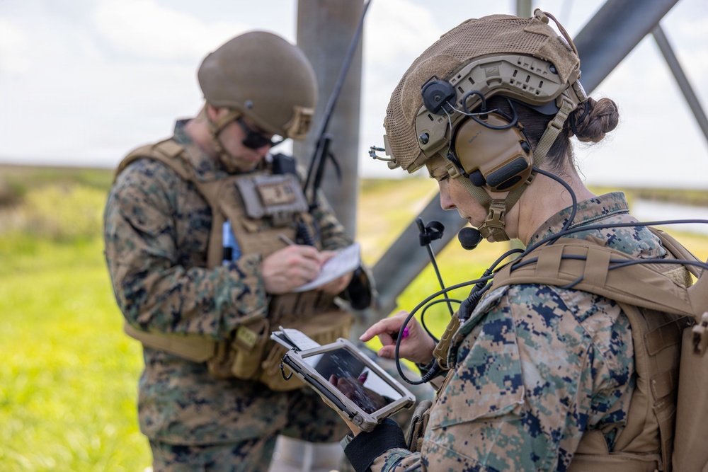 Two military personnel in MARPAT (Marine pattern) camouflage uniforms are engaged in field operations, with one using a ruggedized tablet device and wearing a tactical communications headset with boom microphone. The second individual is writing on a notepad or small document. Both are wearing tactical vests and helmets in an outdoor grassy environment.
