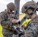 Two military personnel in MARPAT (Marine pattern) camouflage uniforms are engaged in field operations, with one using a ruggedized tablet device and wearing a tactical communications headset with boom microphone. The second individual is writing on a notepad or small document. Both are wearing tactical vests and helmets in an outdoor grassy environment.