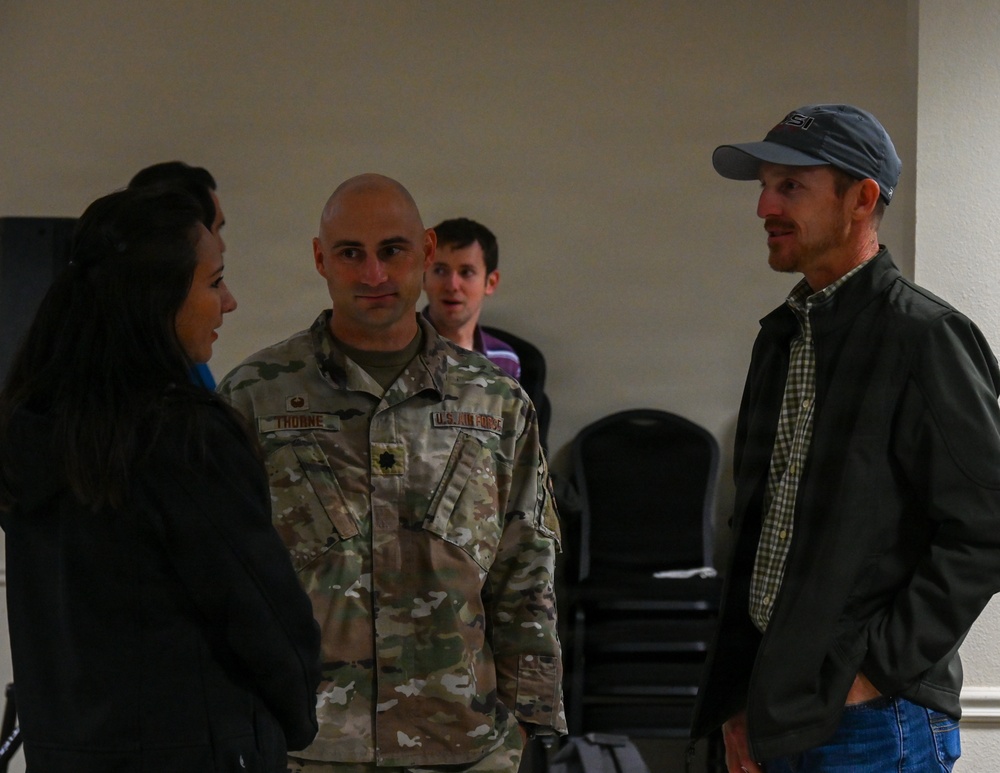 A small group of people are engaged in conversation in what appears to be an indoor meeting or event space. A U.S. Air Force service member in OCP (Operational Camouflage Pattern) uniform with the name tape 'THORNE' and a rank insignia is speaking with a woman in dark clothing and a man in a baseball cap and casual jacket. Additional individuals are visible in the background near stacked chairs.