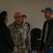 A small group of people are engaged in conversation in what appears to be an indoor meeting or event space. A U.S. Air Force service member in OCP (Operational Camouflage Pattern) uniform with the name tape 'THORNE' and a rank insignia is speaking with a woman in dark clothing and a man in a baseball cap and casual jacket. Additional individuals are visible in the background near stacked chairs.