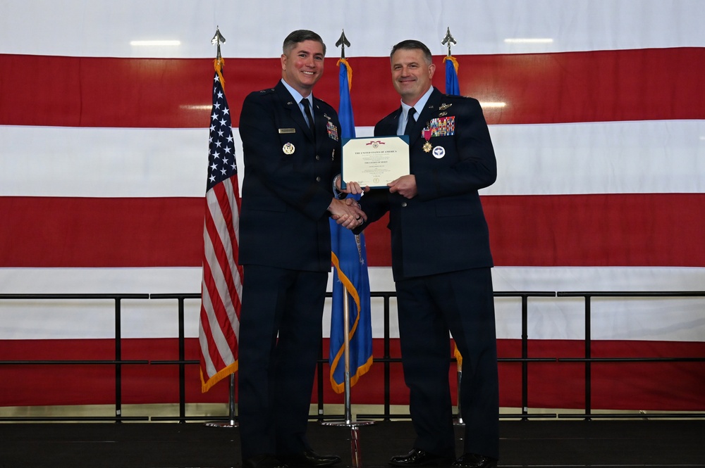 Two U.S. Air Force officers in dress blue uniforms are shaking hands while jointly holding a certificate during a formal military ceremony. The setting features a large American flag backdrop along with what appears to be a state or unit flag. The officer on the right is wearing multiple medals and decorations, suggesting this is an awards or retirement ceremony.