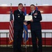 Two U.S. Air Force officers in dress blue uniforms are shaking hands while jointly holding a certificate during a formal military ceremony. The setting features a large American flag backdrop along with what appears to be a state or unit flag. The officer on the right is wearing multiple medals and decorations, suggesting this is an awards or retirement ceremony.
