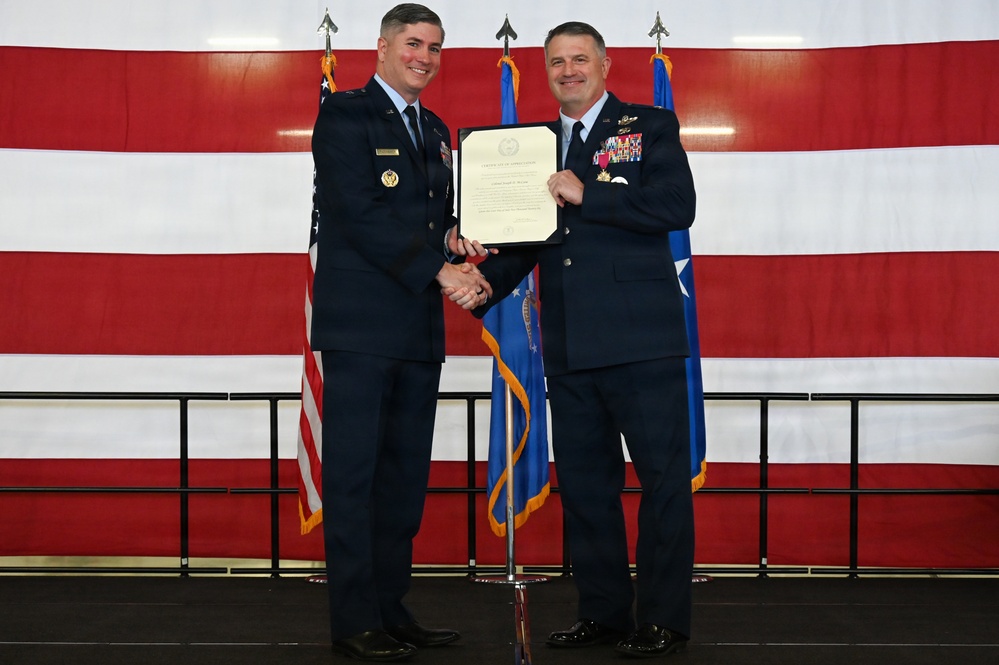 Two U.S. Air Force officers in dress blue uniforms are shaking hands while jointly holding a framed certificate or letter of appreciation during a formal military ceremony. The event appears to be an awards or recognition ceremony, likely a promotion, retirement, or commendation presentation. The backdrop features a large American flag and Air Force flags on a stage.
