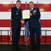 Two U.S. Air Force officers in dress blue uniforms are shaking hands while jointly holding a framed certificate or letter of appreciation during a formal military ceremony. The event appears to be an awards or recognition ceremony, likely a promotion, retirement, or commendation presentation. The backdrop features a large American flag and Air Force flags on a stage.