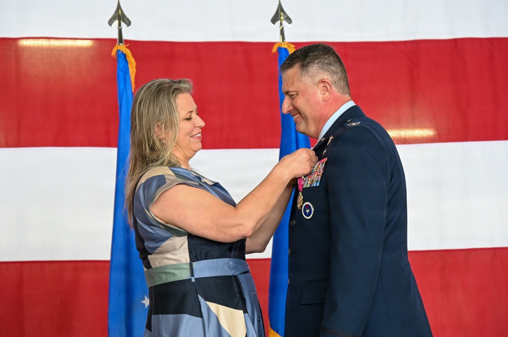 A woman in a patterned dress is pinning a medal onto a male U.S. Air Force officer in dress blues during a formal military ceremony. The officer is wearing multiple service ribbons and medals, and both individuals are smiling. The backdrop features a large American flag and two U.S. Air Force flags on stands.