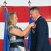 A woman in a patterned dress is pinning a medal onto a male U.S. Air Force officer in dress blues during a formal military ceremony. The officer is wearing multiple service ribbons and medals, and both individuals are smiling. The backdrop features a large American flag and two U.S. Air Force flags on stands.