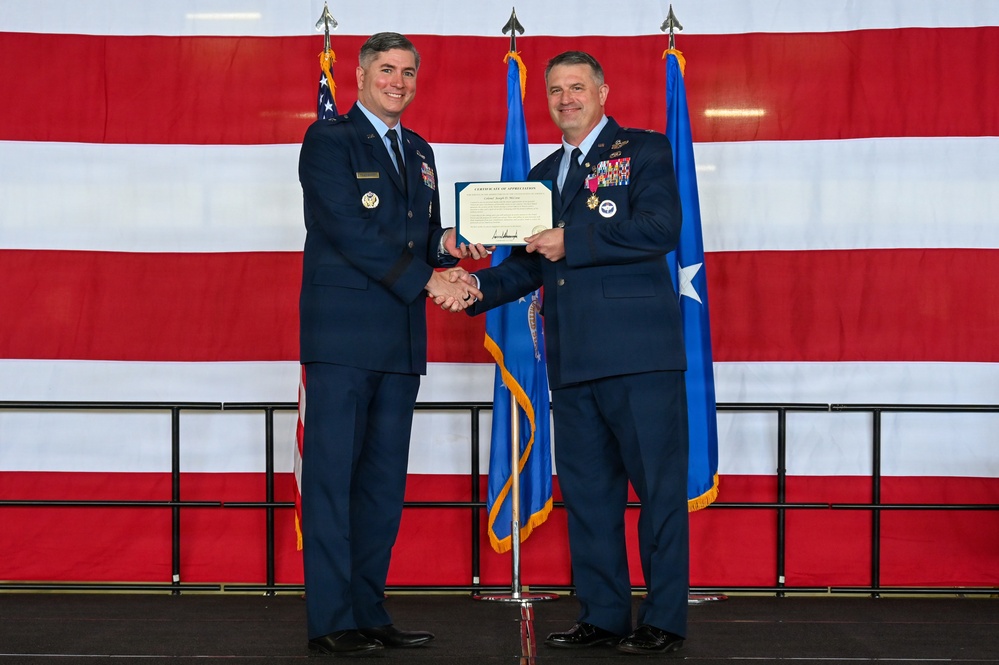 Two U.S. Air Force officers in dress blue uniforms are shaking hands while jointly holding a Certificate of Appreciation during a formal military ceremony. The event takes place in front of a large American flag backdrop with military color guard flags flanking them. The officer on the right displays numerous service ribbons and medals, along with a one-star general insignia.