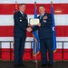 Two U.S. Air Force officers in dress blue uniforms are shaking hands while jointly holding a Certificate of Appreciation during a formal military ceremony. The event takes place in front of a large American flag backdrop with military color guard flags flanking them. The officer on the right displays numerous service ribbons and medals, along with a one-star general insignia.
