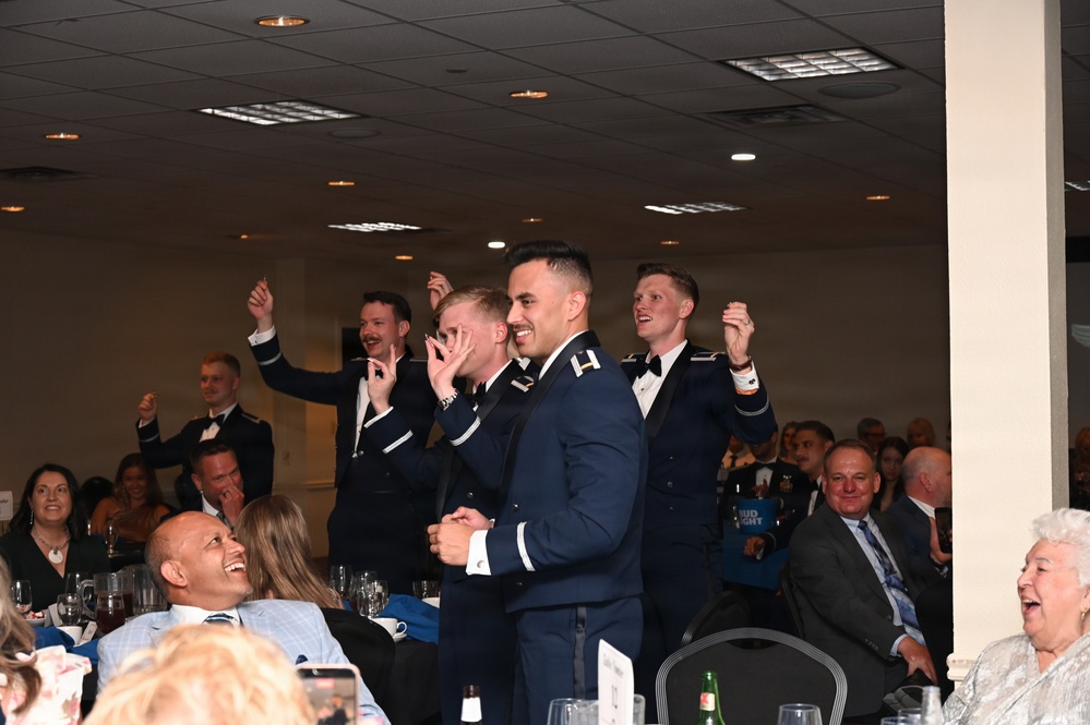 A formal military banquet or ball is taking place in a large event hall. Several uniformed military personnel, appearing to be U.S. Air Force officers in dress blues, are standing and celebrating enthusiastically while seated guests laugh and react around them. The atmosphere is festive and celebratory, suggesting an award ceremony, toast, or special announcement.