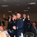 A formal military banquet or ball is taking place in a large event hall. Several uniformed military personnel, appearing to be U.S. Air Force officers in dress blues, are standing and celebrating enthusiastically while seated guests laugh and react around them. The atmosphere is festive and celebratory, suggesting an award ceremony, toast, or special announcement.