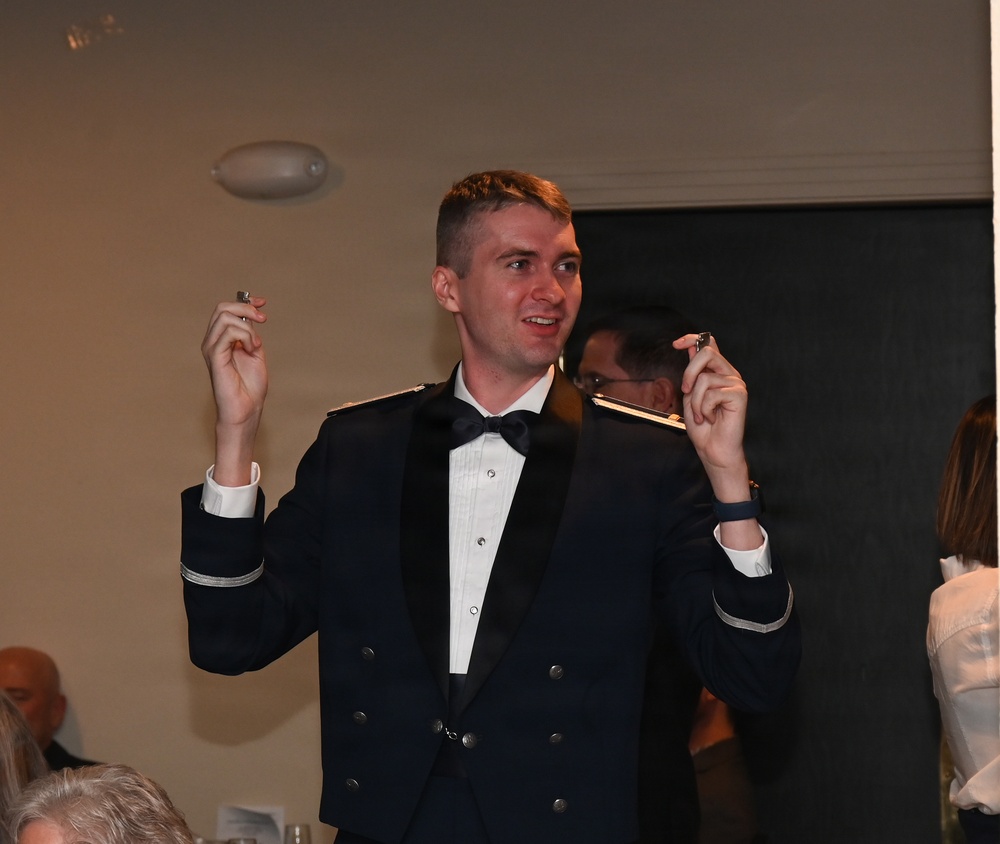 A young man in a dark military dress uniform with a bow tie stands in what appears to be a formal banquet or ceremony setting. He is holding up two small objects, one in each hand, with a slight smile on his face. Other attendees are visible in the background.