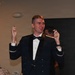 A young man in a dark military dress uniform with a bow tie stands in what appears to be a formal banquet or ceremony setting. He is holding up two small objects, one in each hand, with a slight smile on his face. Other attendees are visible in the background.