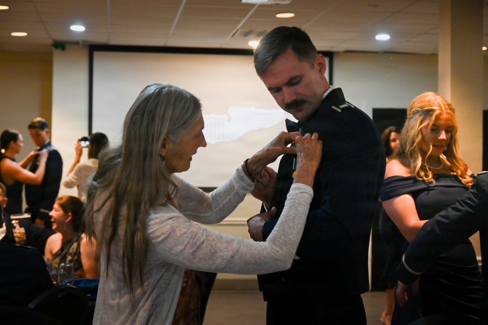 An older woman with long gray hair is pinning something onto the chest of a man wearing a dark military dress uniform. The setting appears to be a formal military ceremony or banquet held in a conference or event hall. Other attendees in formal attire are visible in the background.