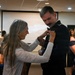An older woman with long gray hair is pinning something onto the chest of a man wearing a dark military dress uniform. The setting appears to be a formal military ceremony or banquet held in a conference or event hall. Other attendees in formal attire are visible in the background.
