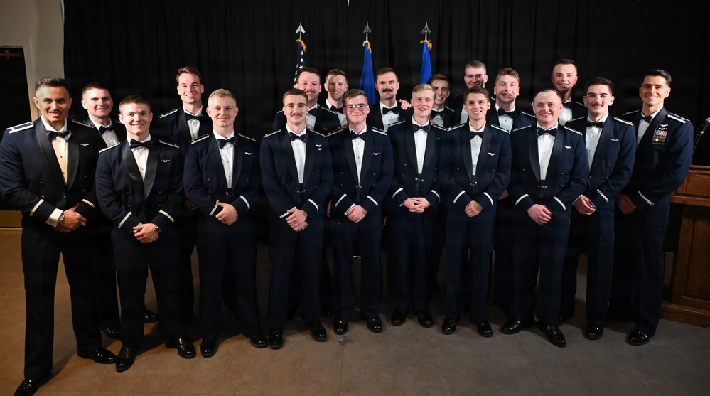 A group of approximately 20 young men in formal U.S. Air Force dress uniforms pose for an official group photograph. They are arranged in two rows against a dark backdrop with American and Air Force flags visible in the background. The occasion appears to be a formal military ceremony or graduation event, indicated by their dress blues and wing insignia.