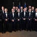 A group of approximately 20 young men in formal U.S. Air Force dress uniforms pose for an official group photograph. They are arranged in two rows against a dark backdrop with American and Air Force flags visible in the background. The occasion appears to be a formal military ceremony or graduation event, indicated by their dress blues and wing insignia.