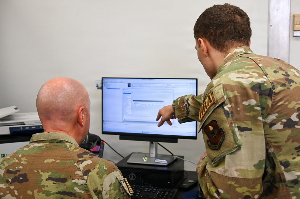 Two military personnel in OCP (Operational Camouflage Pattern) uniforms are reviewing content on a computer monitor. One individual is seated while the other stands and points at the screen, suggesting a collaborative work or training session. A unit patch and name tape are visible on the standing individual's uniform.