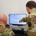 Two military personnel in OCP (Operational Camouflage Pattern) uniforms are reviewing content on a computer monitor. One individual is seated while the other stands and points at the screen, suggesting a collaborative work or training session. A unit patch and name tape are visible on the standing individual's uniform.