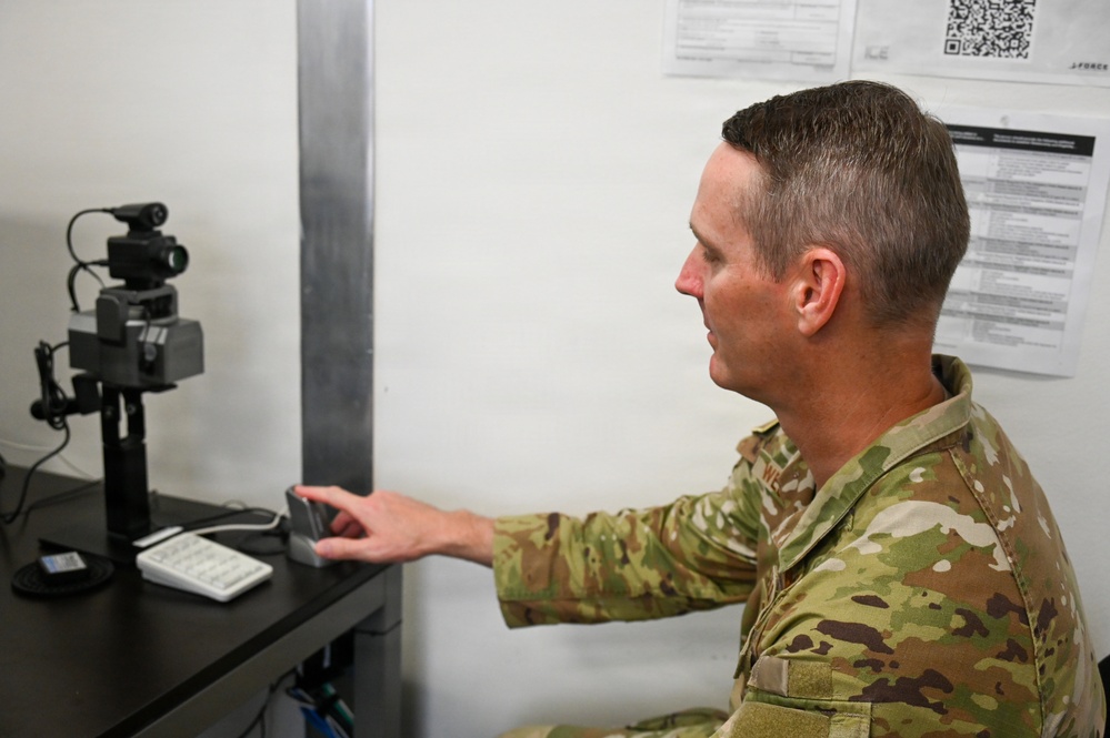 A military service member in OCP (Operational Camouflage Pattern) uniform is seated at a desk using what appears to be a biometric fingerprint scanner. The setting appears to be an administrative or processing station, with documents and a QR code posted on the wall behind him. A camera or optical device is mounted on a stand to his left, suggesting a biometric enrollment or identity verification process.