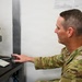 A military service member in OCP (Operational Camouflage Pattern) uniform is seated at a desk using what appears to be a biometric fingerprint scanner. The setting appears to be an administrative or processing station, with documents and a QR code posted on the wall behind him. A camera or optical device is mounted on a stand to his left, suggesting a biometric enrollment or identity verification process.