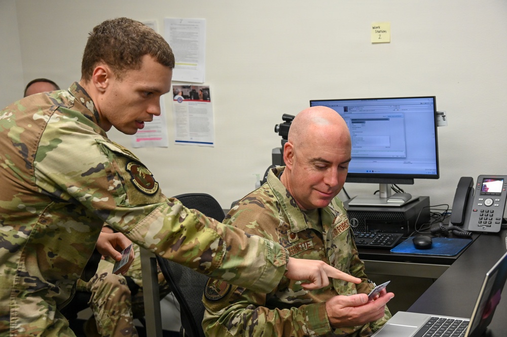 Two military personnel in OCP (Operational Camouflage Pattern) uniforms are working together at a desk workstation. The standing airman appears to be pointing at something on a card or small device held by the seated airman, suggesting a briefing or instructional interaction. The setting appears to be a military office or operations center.