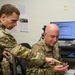 Two military personnel in OCP (Operational Camouflage Pattern) uniforms are working together at a desk workstation. The standing airman appears to be pointing at something on a card or small device held by the seated airman, suggesting a briefing or instructional interaction. The setting appears to be a military office or operations center.