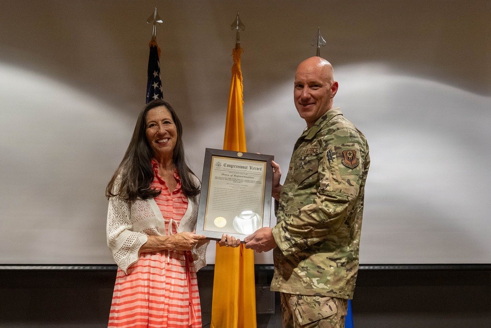 A woman in a pink striped dress and white cardigan and a U.S. Air Force military officer in OCP (Operational Camouflage Pattern) uniform are jointly holding a framed Congressional Record certificate from the House of Representatives. The ceremony appears to be a formal recognition or award presentation. Three flagpoles are visible in the background, including a U.S. flag and what appears to be a New Mexico state flag.