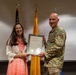 A woman in a pink striped dress and white cardigan and a U.S. Air Force military officer in OCP (Operational Camouflage Pattern) uniform are jointly holding a framed Congressional Record certificate from the House of Representatives. The ceremony appears to be a formal recognition or award presentation. Three flagpoles are visible in the background, including a U.S. flag and what appears to be a New Mexico state flag.
