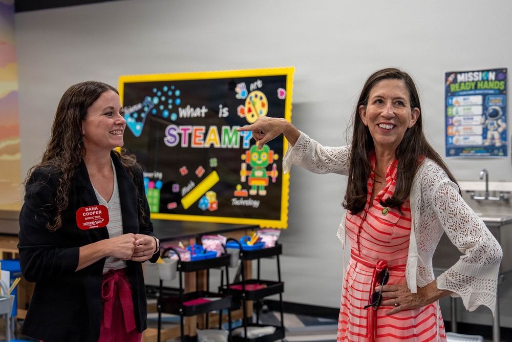 Two women are standing in what appears to be a STEAM education classroom or learning center. The woman on the left wears a name badge identifying her as 'Dana Cooper, Director,' while the woman on the right in a red-striped dress is pointing enthusiastically at something off-frame. Both women are smiling and appear to be engaged in a tour or presentation of the facility.