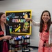Two women are standing in what appears to be a STEAM education classroom or learning center. The woman on the left wears a name badge identifying her as 'Dana Cooper, Director,' while the woman on the right in a red-striped dress is pointing enthusiastically at something off-frame. Both women are smiling and appear to be engaged in a tour or presentation of the facility.