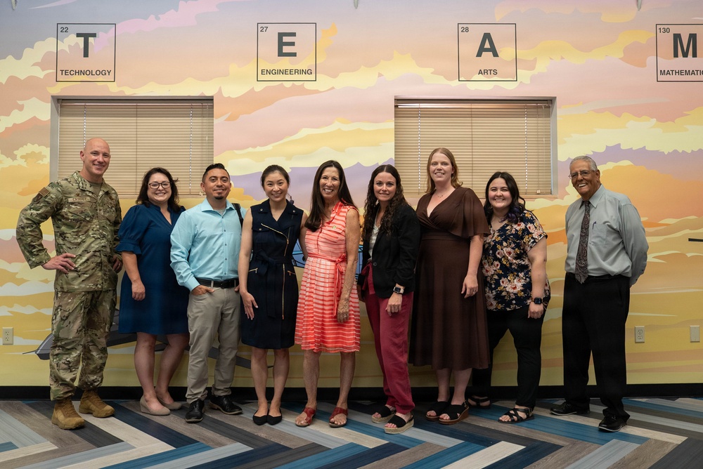 A group of nine adults poses for a formal group photo in front of a colorful STEAM-themed mural. The mural displays periodic table-style tiles spelling out 'TEAM' with the words Technology, Engineering, Arts, and Mathematics. One individual on the far left is wearing military camouflage attire, while the others are dressed in business casual clothing.