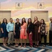 A group of nine adults poses for a formal group photo in front of a colorful STEAM-themed mural. The mural displays periodic table-style tiles spelling out 'TEAM' with the words Technology, Engineering, Arts, and Mathematics. One individual on the far left is wearing military camouflage attire, while the others are dressed in business casual clothing.