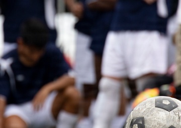 A heavily worn and dirty soccer ball sits in sharp focus on a grass field in the foreground, while a group of players and what appears to be a coach or adult gather in the blurred background. The players are dressed in dark blue and white uniforms with white socks, suggesting a team huddle or break during a match or practice session. The depth-of-field technique draws attention to the weathered ball as the central subject.