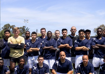A soccer team poses for a formal group photo on an outdoor turf field under a partly cloudy sky. The majority of players are wearing matching navy blue and white uniforms, with two individuals in non-uniform attire likely serving as coaches or staff. The group is arranged in two rows — a standing back row and a crouching front row — in a classic team portrait formation.