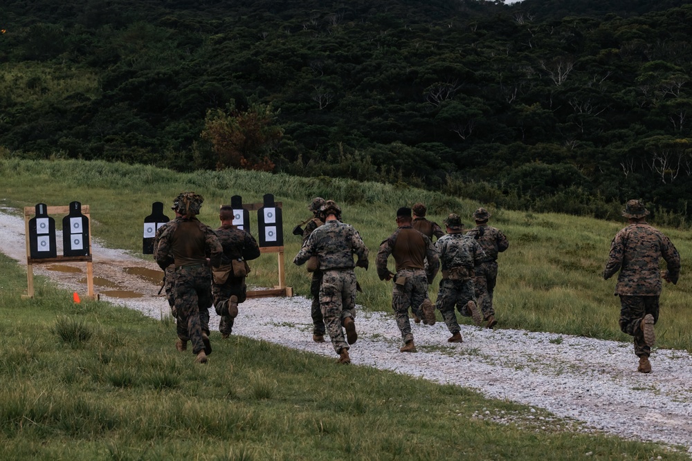 A group of military personnel in camouflage uniforms are running along a gravel path at what appears to be an outdoor shooting range. Multiple paper silhouette targets on wooden stands are visible along the left side of the path. The setting appears to be a training exercise conducted in a lush, green, forested environment.