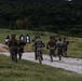 A group of military personnel in camouflage uniforms are running along a gravel path at what appears to be an outdoor shooting range. Multiple paper silhouette targets on wooden stands are visible along the left side of the path. The setting appears to be a training exercise conducted in a lush, green, forested environment.