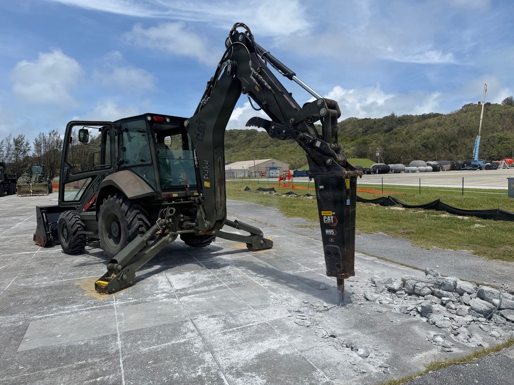 A dark olive/military-colored Caterpillar backhoe loader is actively breaking concrete pavement using a CAT H95 hydraulic hammer attachment. The machine is stabilized with outrigger legs deployed on a concrete surface, with broken concrete rubble visible at the point of impact. The setting appears to be a military installation or airfield, with storage tanks, buildings, and green hills visible in the background.