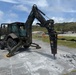 A dark olive/military-colored Caterpillar backhoe loader is actively breaking concrete pavement using a CAT H95 hydraulic hammer attachment. The machine is stabilized with outrigger legs deployed on a concrete surface, with broken concrete rubble visible at the point of impact. The setting appears to be a military installation or airfield, with storage tanks, buildings, and green hills visible in the background.
