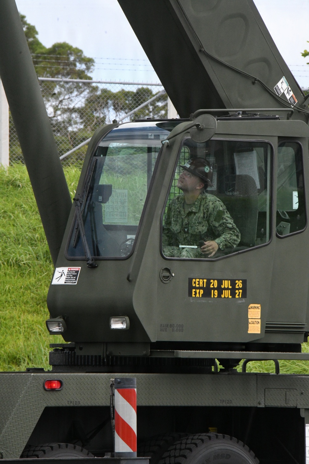 A military service member in camouflage uniform is seated in the cab of an olive-drab military crane vehicle, looking upward, likely monitoring a lifting operation. The vehicle displays a certification placard reading 'CERT 20 JUL 26 / EXP 19 JUL 27', indicating current equipment certification status. The setting appears to be a military installation with a chain-link fence and vegetation visible in the background.