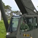 A military service member in camouflage uniform is seated in the cab of an olive-drab military crane vehicle, looking upward, likely monitoring a lifting operation. The vehicle displays a certification placard reading 'CERT 20 JUL 26 / EXP 19 JUL 27', indicating current equipment certification status. The setting appears to be a military installation with a chain-link fence and vegetation visible in the background.