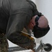A worker is using a yellow angle grinder to cut through metal, producing a shower of bright orange sparks. The person is bent over the workpiece near a white stucco wall, wearing protective safety glasses and ear protection. The cutting operation appears to involve a metal frame or bracket with caster wheels.