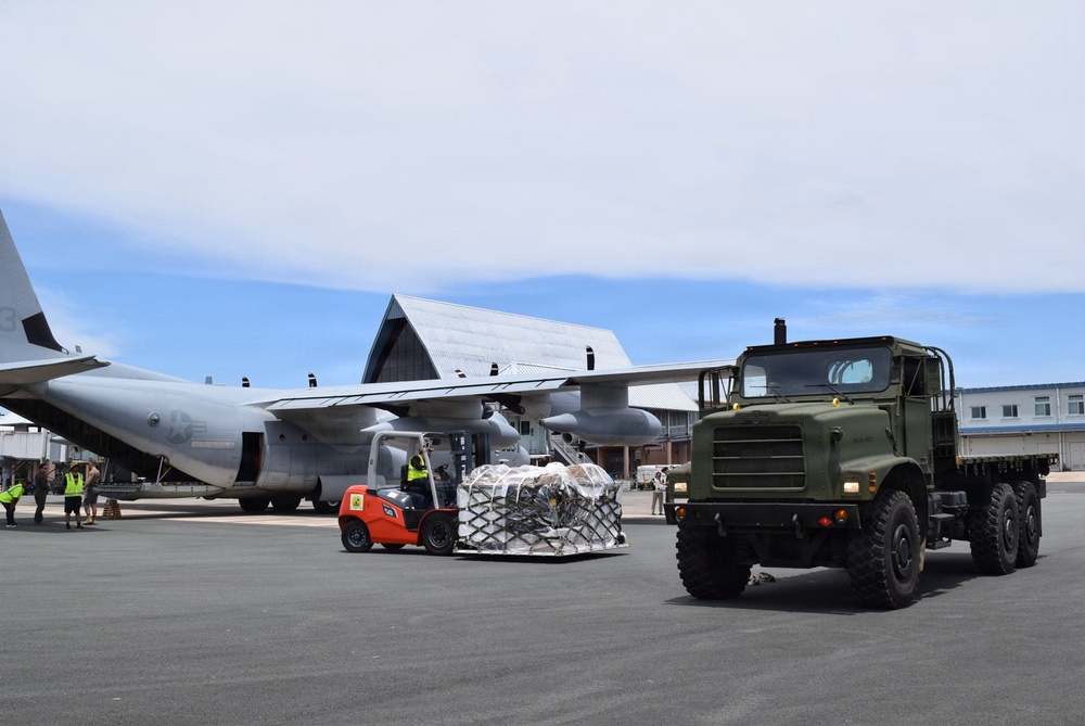 A military cargo offloading operation is underway on an airport tarmac. A C-130 Hercules transport aircraft is being unloaded using an orange forklift carrying a netted cargo pallet, while a green military 6x6 cargo truck stands nearby. Ground personnel in high-visibility vests are visible coordinating the operation near a terminal building.