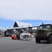 A military cargo offloading operation is underway on an airport tarmac. A C-130 Hercules transport aircraft is being unloaded using an orange forklift carrying a netted cargo pallet, while a green military 6x6 cargo truck stands nearby. Ground personnel in high-visibility vests are visible coordinating the operation near a terminal building.