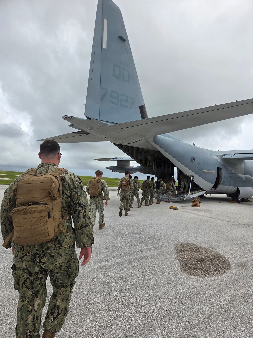 Military personnel in camouflage uniforms are boarding a large gray military transport aircraft via its rear ramp. The aircraft's tail markings show 'QD' and the number '7927', consistent with a C-130 Hercules-type transport. Personnel are carrying tactical backpacks and appear to be loading onto the aircraft on an open tarmac under overcast skies.