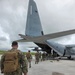 Military personnel in camouflage uniforms are boarding a large gray military transport aircraft via its rear ramp. The aircraft's tail markings show 'QD' and the number '7927', consistent with a C-130 Hercules-type transport. Personnel are carrying tactical backpacks and appear to be loading onto the aircraft on an open tarmac under overcast skies.