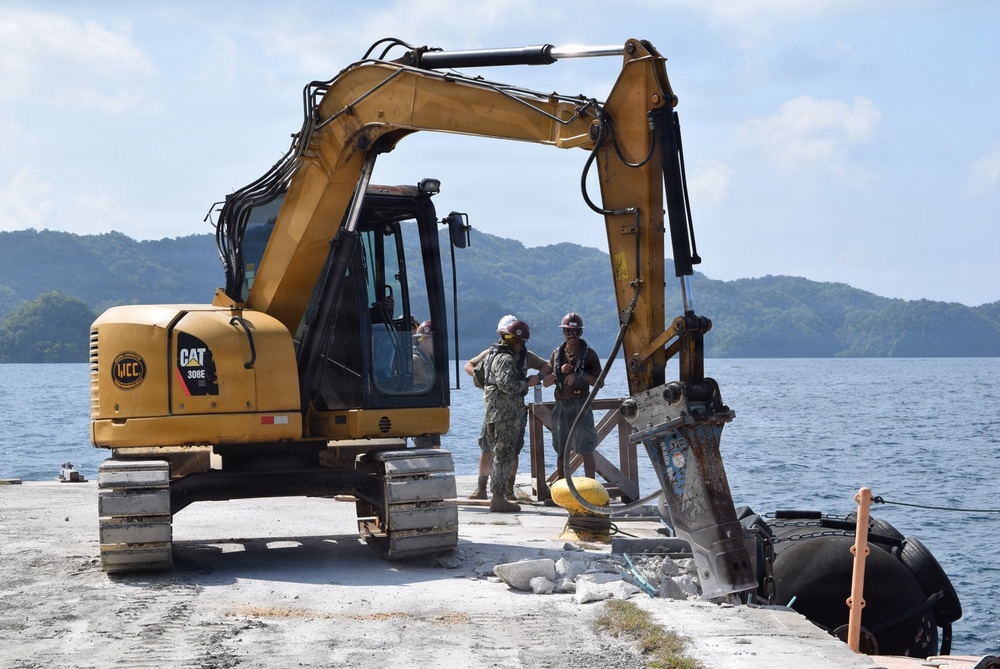 A CAT 308E mini excavator operated by WCC is positioned on a concrete pier or dock near a body of water, equipped with a hydraulic breaker attachment being used for demolition work. Two workers wearing hard hats and military-style camouflage uniforms are standing near the machine, apparently coordinating the operation. The setting appears to be a tropical coastal location with forested hills visible in the background.
