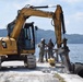 A CAT 308E mini excavator operated by WCC is positioned on a concrete pier or dock near a body of water, equipped with a hydraulic breaker attachment being used for demolition work. Two workers wearing hard hats and military-style camouflage uniforms are standing near the machine, apparently coordinating the operation. The setting appears to be a tropical coastal location with forested hills visible in the background.