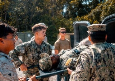 Military personnel in camouflage uniforms are carrying an individual on a stretcher or litter, suggesting a medical evacuation or casualty training exercise. Multiple service members are involved in the carry, with additional personnel observing in the background. The setting appears to be an outdoor military installation or training area.