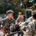 Military personnel in camouflage uniforms are carrying an individual on a stretcher or litter, suggesting a medical evacuation or casualty training exercise. Multiple service members are involved in the carry, with additional personnel observing in the background. The setting appears to be an outdoor military installation or training area.